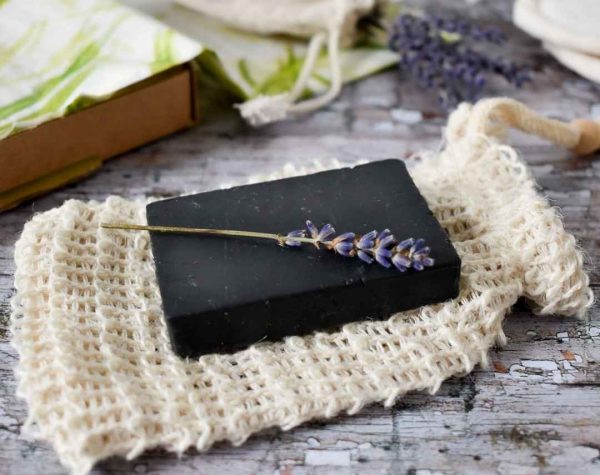 white A black soap bar and purple lavender sprig are placed on top of a beige soap saver bag . Background is a grey wooden table.