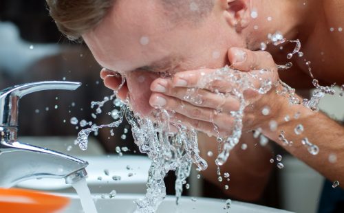 Man washing face over sink with faucet running